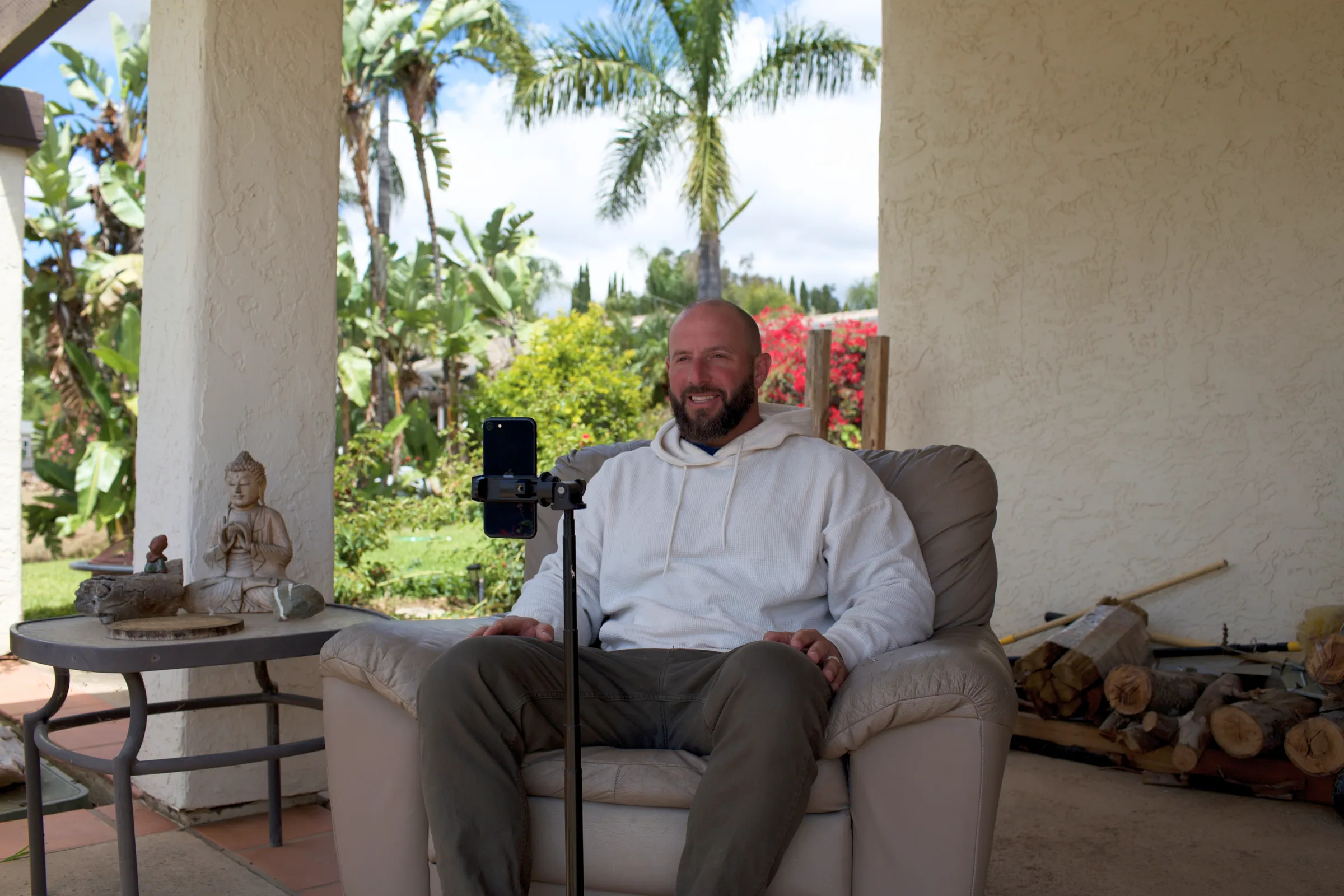 Miles sitting in an armchair on a covered porch, smiling toward a phone on a tripod