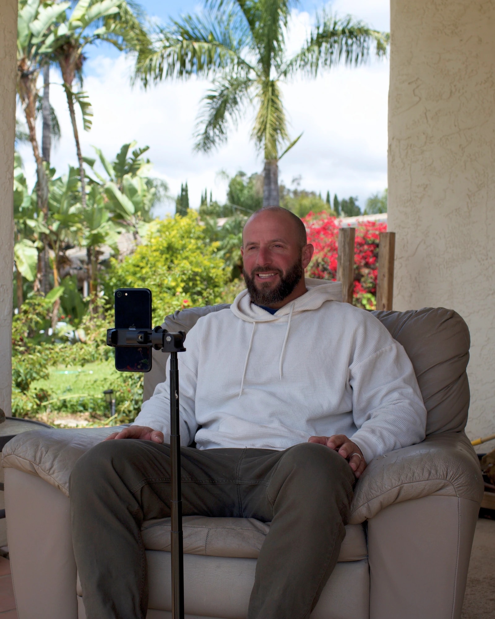 Miles sitting in an armchair on a covered porch, smiling toward a phone on a tripod